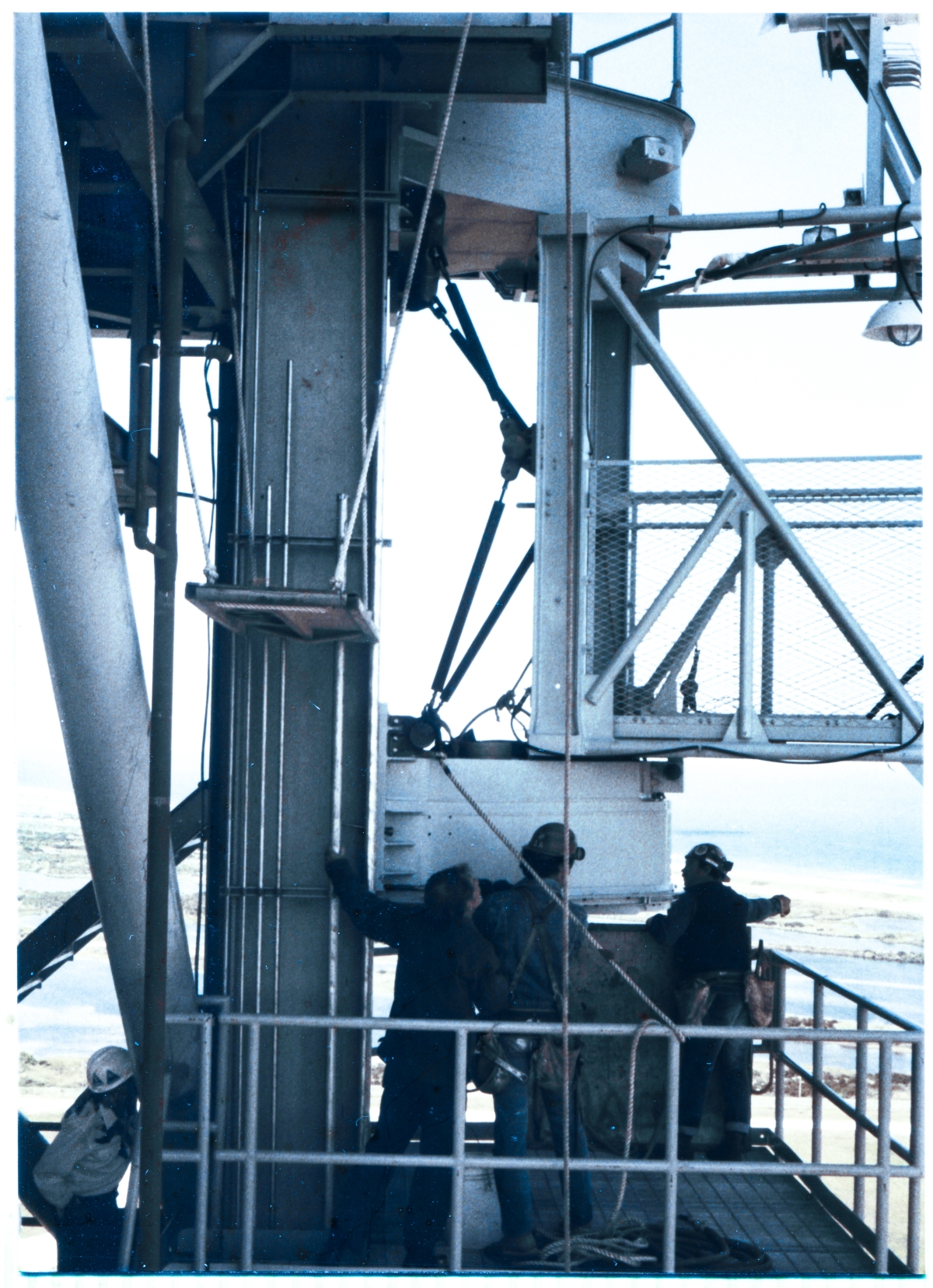 Image 116. Work on attaching the GOX Arm to the Fixed Service Structure at Space Shuttle Launch Complex 39-B, Kennedy Space Center, Florida, by Union Ironworkers from Local 808 working for Ivey Steel continues, over 250 above the distant wilderness of the Merritt Island Wildlife Refuge visible in the background, with very fine adjustments in the positioning and orientation of the Arm's Hinge Boxes up against the Strongback Columns on the FSS that they will be bolted to, ongoing. The near side of the Lower Hinge Box appears to be in snug contact with the Strongback, and one of the ironworkers has driven a bull pin through one of the matched hole-sets between Arm and Strongback there, restraining it from further horizontal movement at the point of insertion. Above, the Upper Hinge Box remains just out of contact with the Strongback, and the TT&V guy without a hardhat, left hand on a conduit, surveys the situation in between spells of giving instructions to the Crane Operator via walkie-talkie. At the far corner of the Lower Hinge Access Platform, one of the ironworkers is giving hand signals, and it appears as if the GOX Arm needs to be raised, just a bit, to bring the Upper Hinge Box into snug contact with the Strongback. In the lower left corner of the frame, one of the TT&V QC people observing the work looks up at the ongoing activities from a safe place. The Arm is where it belongs, but it will be a while yet before all of the attachment bolts can be inserted into their holes and fully torqued-down, fastening the Arm onto the Strongback in its permanent working position on the tower. Photo by James MacLaren.